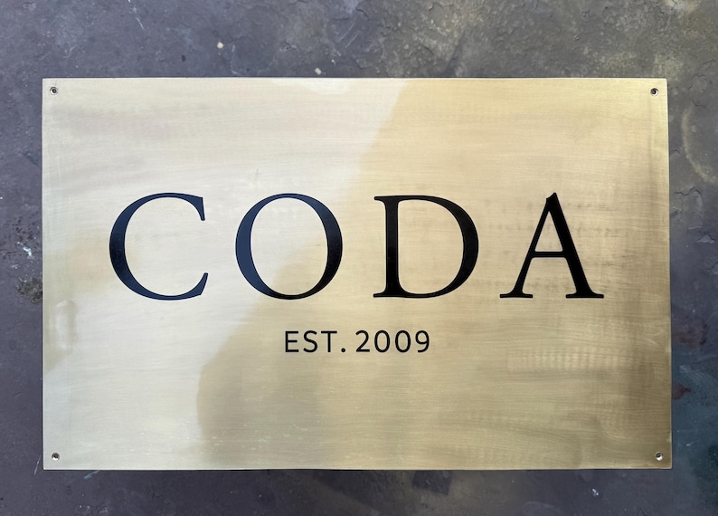May include: A rectangular brass sign with the word "CODA" in bold black letters. Below the word is the text "EST. 2009" in a smaller, black font. The sign has a brushed finish and is mounted with four small screws.