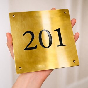 May include: A gold-colored metal sign with the number "201" in black. The sign is square with rounded corners and four screw holes. The sign is being held by a person.