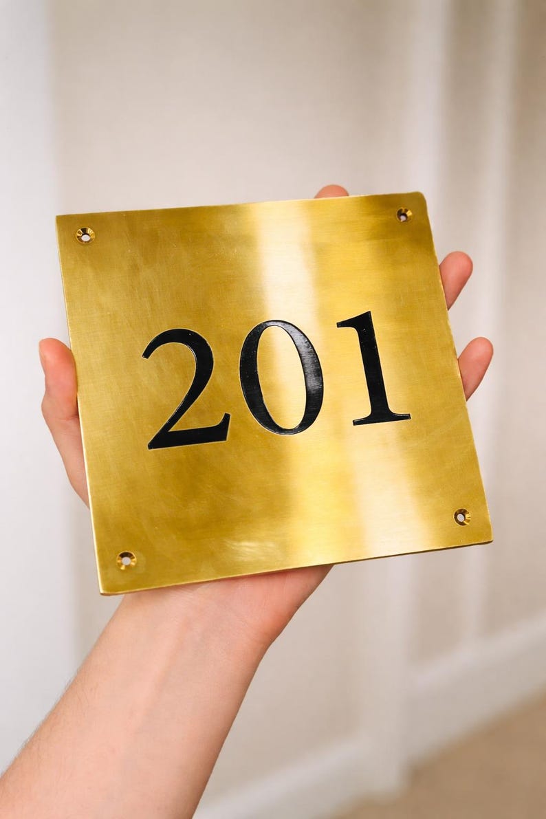 May include: A gold-colored metal sign with the number "201" in black. The sign is square with rounded corners and four screw holes. The sign is being held by a person.