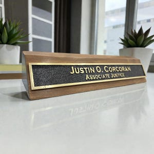 May include: A wooden desk nameplate with a brass-colored metal plate. The plate has the name "JUSTIN O. CORCORAN" and the title "ASSOCIATE JUSTICE" in gold lettering. The nameplate is rectangular and sits on a white surface.