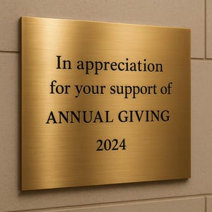May include: A rectangular, brushed gold plaque with black text. The text reads "In appreciation for your support of ANNUAL GIVING 2024". The plaque is mounted on a light-colored, textured wall.