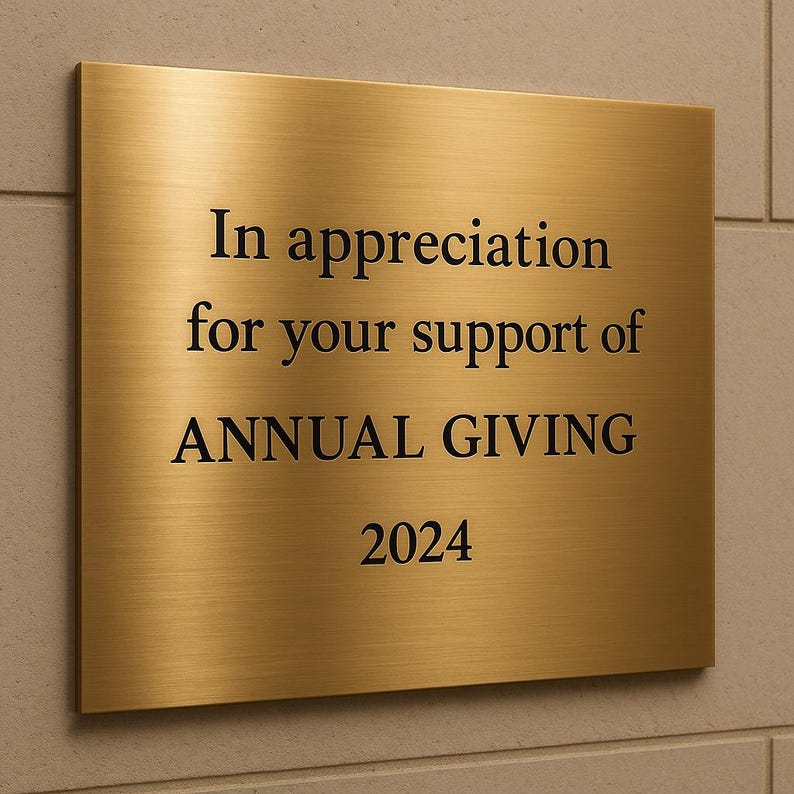 May include: A rectangular, brushed gold plaque with black text. The text reads "In appreciation for your support of ANNUAL GIVING 2024". The plaque is mounted on a light-colored, textured wall.