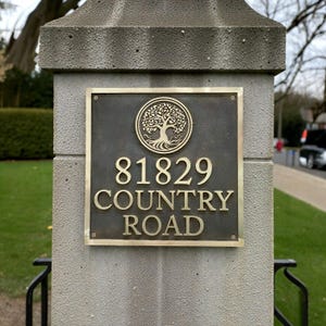 May include: A square bronze address plaque mounted on a gray stone pillar. The plaque features a circular emblem of a tree at the top, followed by the numbers 81829 and the text COUNTRY ROAD in raised, polished gold-toned lettering.