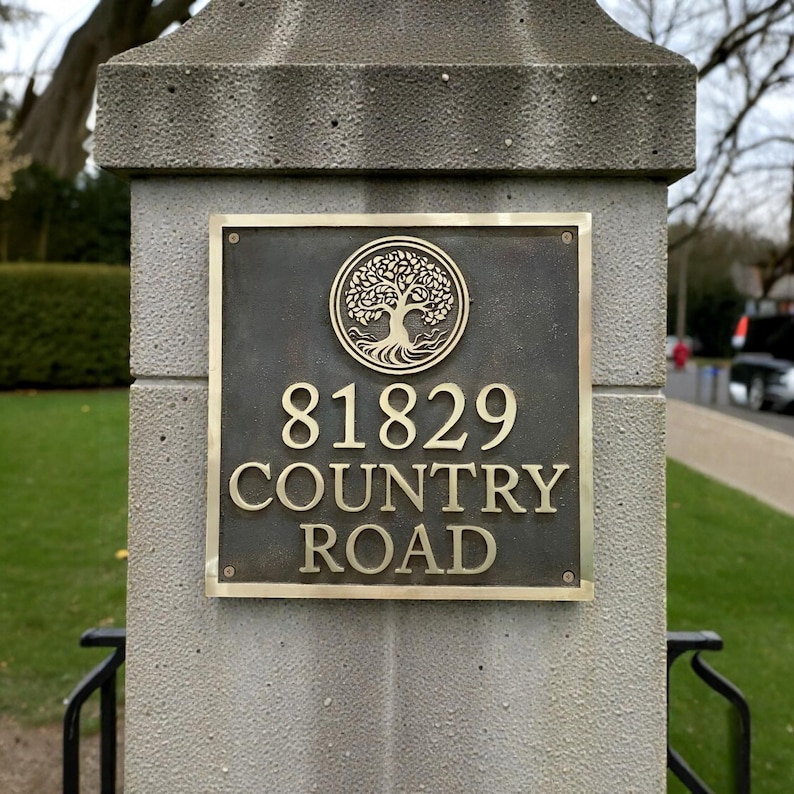 May include: A square bronze address plaque mounted on a gray stone pillar. The plaque features a circular emblem of a tree at the top, followed by the numbers 81829 and the text COUNTRY ROAD in raised, polished gold-toned lettering.