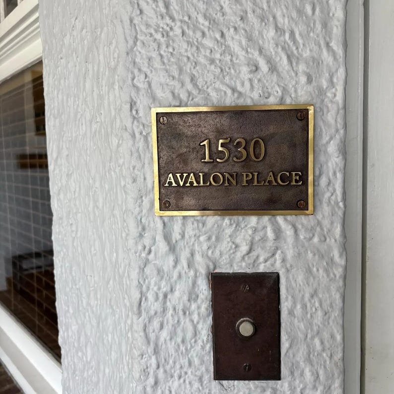May include: A rectangular bronze-toned metal address plaque featuring the number 1530 and the text AVALON PLACE in raised gold-colored lettering, secured with four screws, positioned above a matching rectangular doorbell button on a white wall.