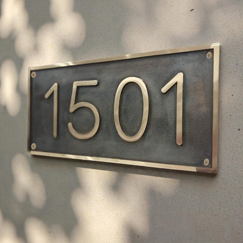 May include: A rectangular house number plaque featuring the digits 1501 in a metallic finish. The plaque has a dark, textured gray center panel surrounded by a polished metallic frame, secured to a wall with four visible corner screws.