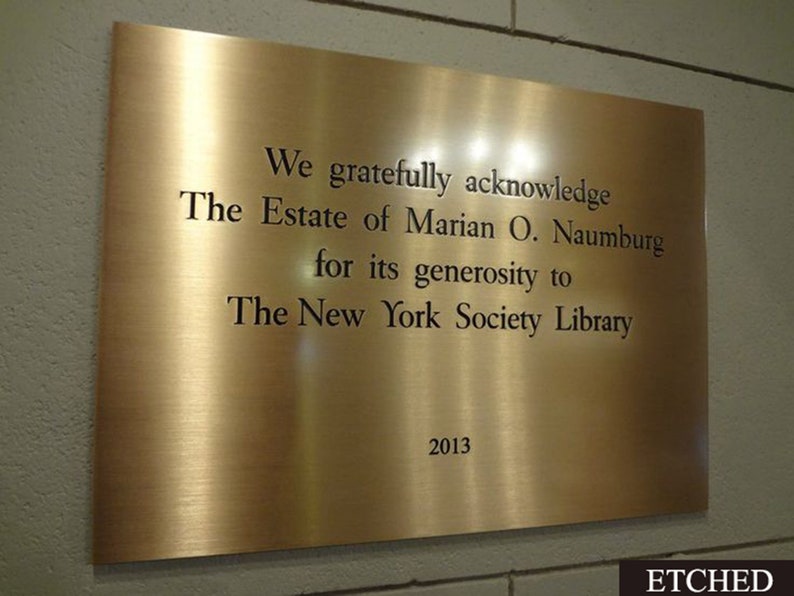 May include: A rectangular, brushed brass plaque with engraved text. The text reads: "We gratefully acknowledge The Estate of Marian O. Naumburg for its generosity to The New York Society Library 2013." The word "ETCHED" is in the bottom right corner.