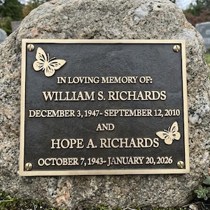 May include: A rectangular bronze memorial plaque mounted on a large stone, featuring the text IN LOVING MEMORY OF: WILLIAM S. RICHARDS DECEMBER 3, 1947- SEPTEMBER 12, 2010 AND HOPE A. RICHARDS OCTOBER 7, 1943- JANUARY 20, 2026 with two butterfly motifs.