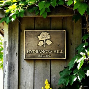 May include: A rectangular bronze-colored metal plaque featuring a raised hydrangea flower graphic at the top, with the text HYDRANGEA HILL and EST. 2023 embossed in capital letters below the flower, mounted on a vertical wooden surface.