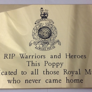 May include: A rectangular, gold-colored plaque with engraved text and a crest. The text reads "RIP Warriors and Heroes" and "This Poppy is dedicated to all those Royal Marines who never came home." The crest includes a globe and the words "Gibraltar" and "Per Mare Per Terram."