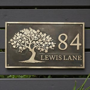 May include: A rectangular metal house sign with a dark, weathered finish featuring a raised brass-colored tree silhouette on the left side. The number 84 is displayed in large, raised brass-colored numerals, with the text LEWIS LANE below.