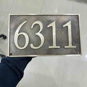 May include: A rectangular house number plaque featuring the digits 6311 in a polished metallic finish. The background has a dark, textured, weathered bronze appearance with a smooth metallic border and four small circular mounting holes.