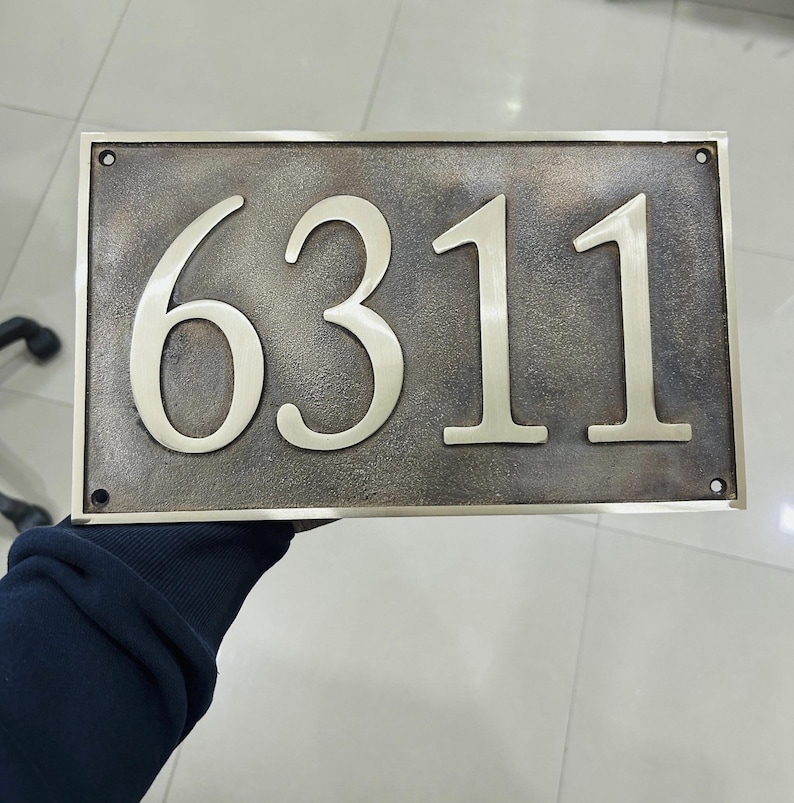 May include: A rectangular house number plaque featuring the digits 6311 in a polished metallic finish. The background has a dark, textured, weathered bronze appearance with a smooth metallic border and four small circular mounting holes.