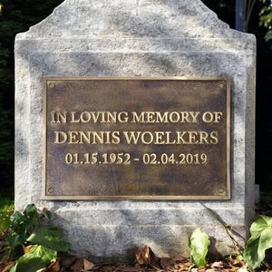 Könnte beinhalten: Ein Steinmonument mit einer Bronzeplatte. Auf der Platte steht in goldener Schrift: "IN LOVING MEMORY OF DENNIS WOELKERS 01.15.1952 - 02.04.2019". Das Denkmal befindet sich in einer grünen Umgebung.