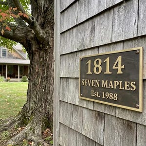 May include: A rectangular bronze-toned metal plaque with a raised gold-colored border and lettering. The plaque displays the number 1814, the text SEVEN MAPLES, and the text Est. 1988, secured with four visible screws at the corners of the metal plate.