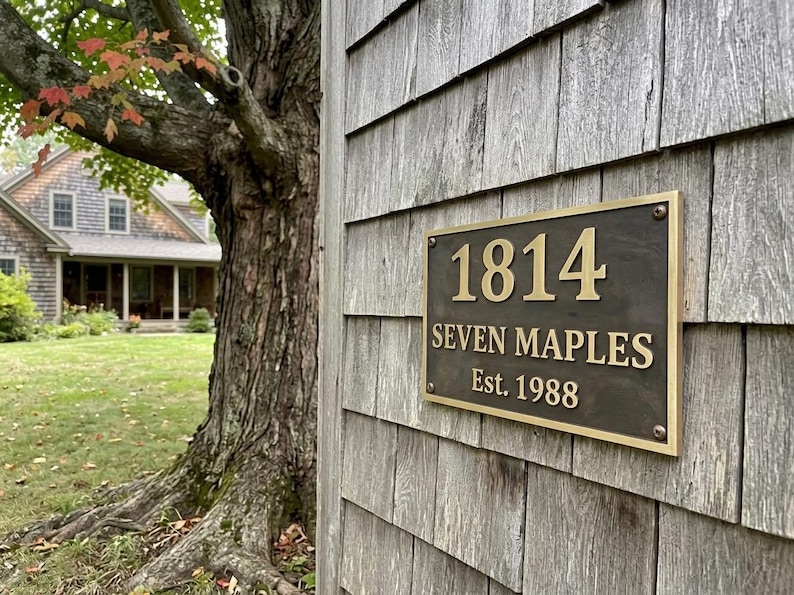 May include: A rectangular bronze-toned metal plaque with a raised gold-colored border and lettering. The plaque displays the number 1814, the text SEVEN MAPLES, and the text Est. 1988, secured with four visible screws at the corners of the metal plate.