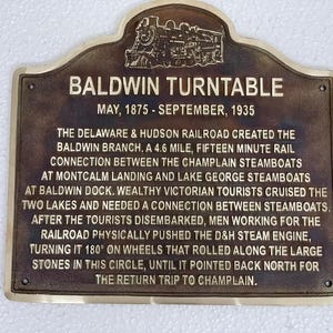 Puede incluir: Una placa de metal color bronce de forma rectangular con la parte superior redondeada y una locomotora de vapor en relieve. El texto dice: BALDWIN TURNTABLE, MAY, 1875 - SEPTEMBER, 1935, seguido de una descripci&oacute;n hist&oacute;rica del ferrocarril.