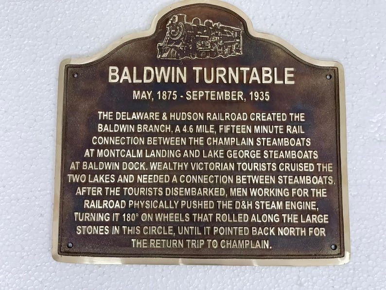 Puede incluir: Una placa de metal color bronce de forma rectangular con la parte superior redondeada y una locomotora de vapor en relieve. El texto dice: BALDWIN TURNTABLE, MAY, 1875 - SEPTEMBER, 1935, seguido de una descripci&oacute;n hist&oacute;rica del ferrocarril.