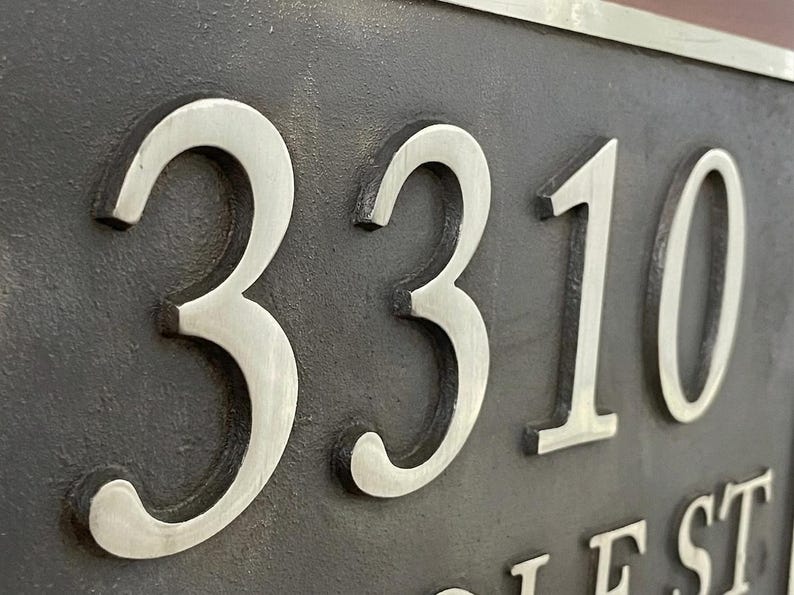 May include: A rectangular house number plaque featuring the raised silver-colored digits 3310 set against a dark, textured charcoal-colored background. The numbers have a smooth, metallic finish and are clearly legible against the matte surface.
