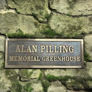 May include: A bronze plaque with the words "ALAN PILLING MEMORIAL GREENHOUSE" in gold lettering is mounted on a weathered stone wall. The wall is composed of large, irregularly shaped stones covered in green moss.