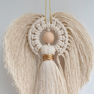 Pode incluir: Um ornamento de anjo de macramé branco com cabeça de madeira e detalhes dourados. O anjo tem cabelos longos e soltos e um vestido longo e solto.
