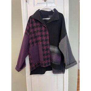 May include: A long-sleeve pullover top featuring a color-blocked design with a black collar and right side, a purple and black houndstooth pattern on the left side, a solid purple sleeve, a grey sleeve, and a purple and grey plaid patch pocket.