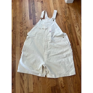 May include: Off-white cotton overalls featuring a bib pocket with a button closure, adjustable shoulder straps with metal buckles, side button closures at the waist, and two front pockets. The garment includes a small rectangular brand label.