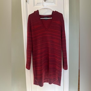 May include: A long-sleeve hooded sweater dress in a deep burgundy and red heathered knit pattern. The garment features a V-neckline, a hood, and a curved hemline. The fabric has a textured appearance with horizontal tonal stripes throughout the piece.