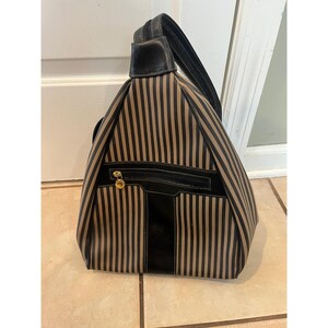 May include: A triangular-shaped backpack featuring a vertical striped pattern in tan and black. The bag includes a black leather-like trim, a single top handle, and a front zippered pocket with a gold-toned circular zipper pull hardware.