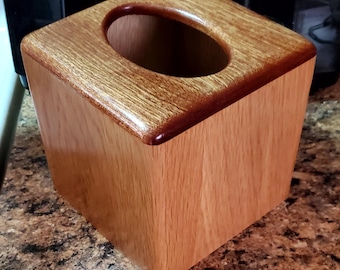 May include: A wooden tissue box with a square shape and a smooth, polished finish. The top features a rounded oval opening for tissue dispensing. The box is made of light brown wood with a darker brown trim around the top edge.