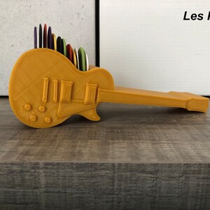 Può includere: Un porta plettri in plastica color oro a forma di chitarra. Il porta plettri ha una superficie testurizzata e il testo "Les Paul" è stampato sullo sfondo bianco dietro il porta plettri.