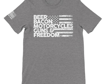 Op de afbeelding: Een grijs t-shirt met een versleten witte afbeelding in de vorm van de Amerikaanse vlag. Het vlagontwerp bevat de woorden "BEER BACON MOTORCYCLES GUNS & FREEDOM". Op de linkermouw staat een kleine Amerikaanse vlag.