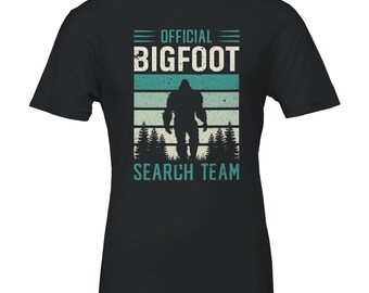 Op de afbeelding: Zwart T-shirt met een Bigfoot-afbeelding. Het ontwerp toont de tekst "OFFICIAL BIGFOOT SEARCH TEAM" in wit en blauwgroen, met een silhouet van Bigfoot die door een bos loopt.