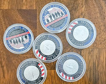 Op de afbeelding: Vijf zilveren challenge-munten met rood-wit-blauwe Amerikaanse vlagontwerpen. De munten bevatten de woorden "Proud of You", "Veterans" en "Thank You" samen met de woorden "Honoring All Who Served" en "For Your Service".