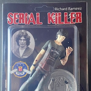 Puede incluir: Una figura de acción de plástico de Richard Ramirez, un asesino en serie, que lleva una camiseta gris con las palabras "Jack Daniels" y una mano ensangrentada. La figura sostiene una pistola en una mano y un cuchillo ensangrentado en la otra. La figura está de pie sobre una base redonda de plata con las palabras "Richard Ramirez" en ella. La figura está empaquetada en una caja de plástico transparente con un fondo negro. La caja tiene las palabras "Serial Killer" en letras rojas en la parte delantera. La caja también tiene una etiqueta de advertencia que dice "ADVERTENCIA: DEFINITIVAMENTE NO PARA NIÑOS".