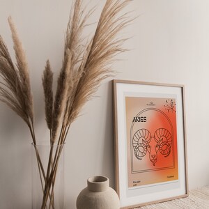 May include: A framed print with a minimalist design featuring the zodiac sign Aries. The print has a gradient background in shades of orange and red. The text "The Pioneer" and "Aries" are in black font. The sign is represented by a ram's head in black outline.