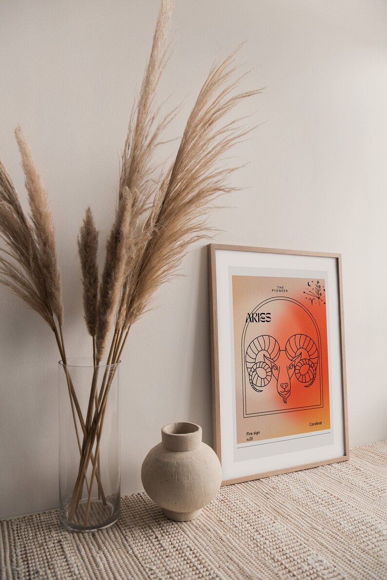 May include: A framed print with a minimalist design featuring the zodiac sign Aries. The print has a gradient background in shades of orange and red. The text "The Pioneer" and "Aries" are in black font. The sign is represented by a ram's head in black outline.