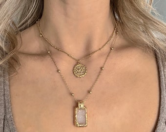 May include: Two gold necklaces with pendants. The top necklace has a round pendant with an intricate design. The bottom necklace has a rectangular pendant with a clear stone inside.