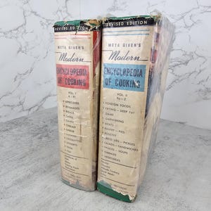 May include: Two volumes of Meta Given's Modern Encyclopedia of Cooking, Revised Edition, wrapped in clear plastic. Volume I features a red spine label with A-Fi, while Volume II features a blue spine label with Fa-Z, both listing various food categories.