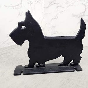 May include: Black cast iron dog-shaped bookend. The silhouette of a Scottish Terrier is depicted in a standing pose, with a rectangular base. The bookend is designed to hold books upright on a shelf or desk.