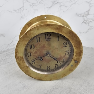 May include: A vintage brass clock with a round face and a weathered appearance. The clock face features black numerals and hands, with a glass cover. The clock's brass casing has a circular design with a mounting hole at the top.