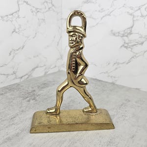 May include: A brass figurine of a man in a military-style uniform, walking with one leg forward. The figurine is on a rectangular base and has a loop at the top. The color is gold.