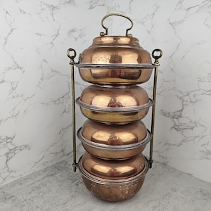 May include: A vintage copper food carrier with four stacked, lidded bowls. The copper has a warm, reddish-brown hue and is supported by a brass-colored metal frame with a handle on top. The bowls are approximately 6 inches in diameter.