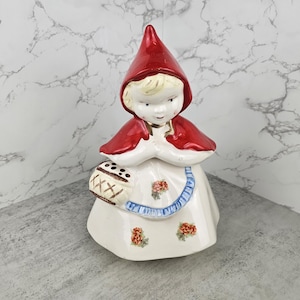 May include: A ceramic figurine of Little Red Riding Hood, featuring a red cape and hood, a white dress with floral accents, and a basket. The figurine has blonde hair and is holding her hands together. The overall design is vintage.