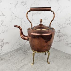 May include: A polished copper kettle featuring a curved spout, a removable lid with a small finial, and a sturdy handle. The kettle is supported by three slender brass legs that elevate the main body, showcasing a metallic finish throughout.