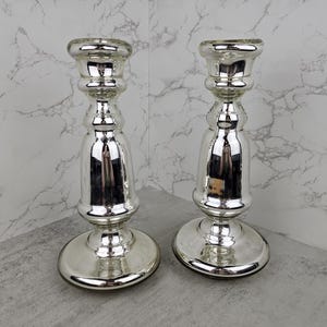 May include: Two silver-colored candle holders with a reflective finish. The candle holders have a round base, a bulbous midsection, and a flared top. They are placed on a gray surface against a marble-patterned background.