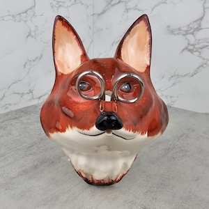 May include: A ceramic fox head wall mount featuring a glossy finish in shades of reddish-brown and white. The fox has pointed ears, a black nose, and a pair of silver-toned metal spectacles resting on its snout, creating a distinct facial feature.