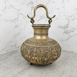 May include: A brass vessel with a rounded body and a curved handle. The surface features intricate embossed relief carvings of figures and floral motifs. The neck has a ribbed texture, and the base rests on small, rounded feet, showcasing a metallic finish.