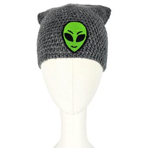Op de afbeelding: Een grijze gebreide beanie met een groene alien gezicht patch. De beanie heeft kleine oorachtige punten aan de bovenkant. De alien patch heeft een groen gezicht met zwarte ogen en een lachende mond. De hoed wordt getoond op een witte mannequin hoofd.