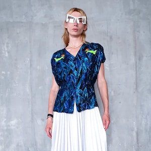 Op de afbeelding: Een blauw patroon top met een V-hals en korte mouwen. De top heeft een zwart-blauw ontwerp met twee groene geborduurde figuren. De top wordt gecombineerd met een witte geplooide rok. Het model draagt een witte zonnebril.